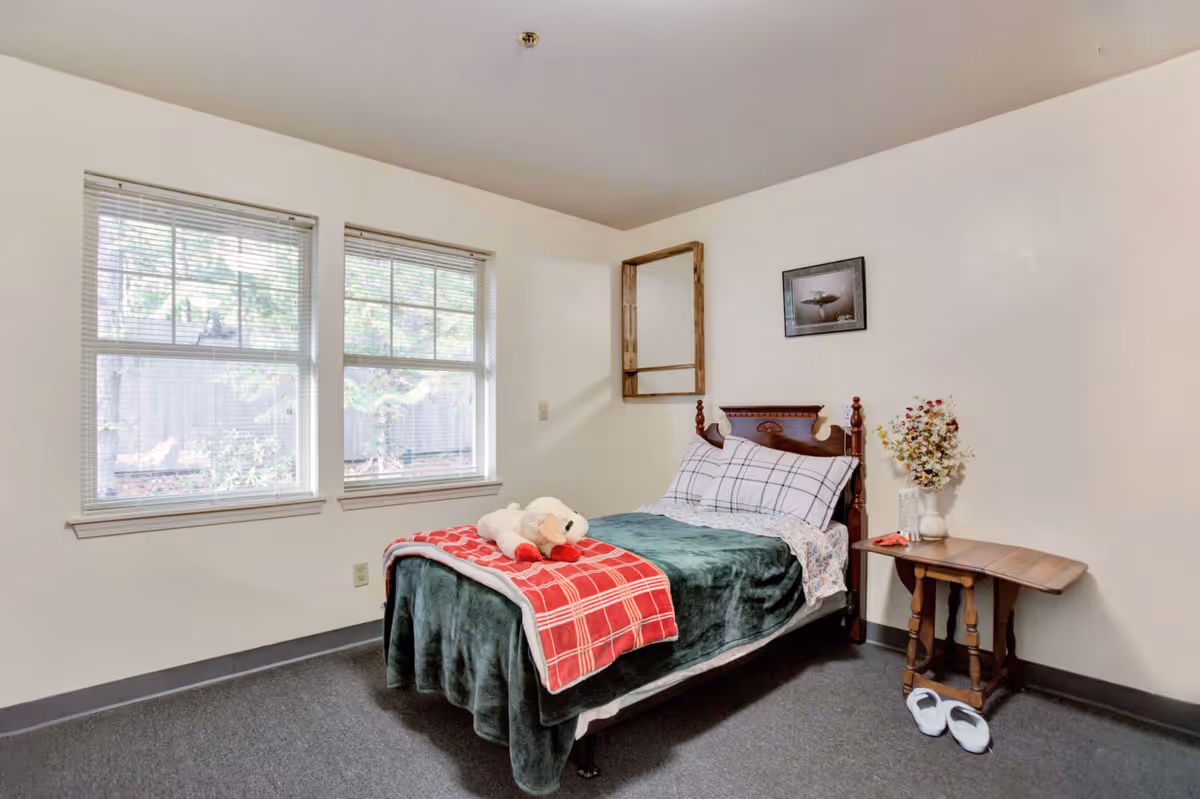A simple bedroom with a single bed covered with a green blanket and a red plaid throw. A stuffed animal is placed on the bed. There are two windows with blinds on the left wall, a small wooden side table with a vase of flowers, and a pair of white slippers on the floor. A framed picture hangs on the wall above the bed.
