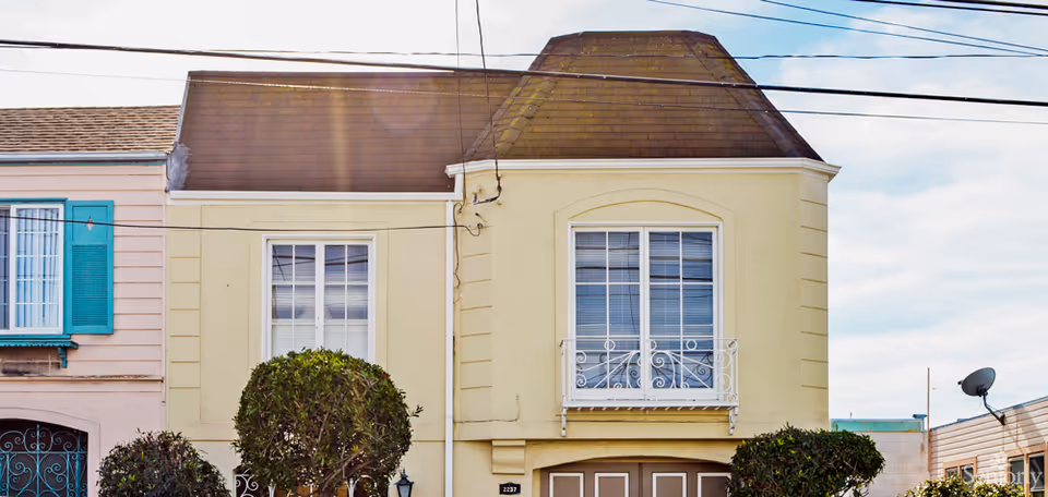 Front exterior view of a two-story residential building with a beige facade, brown roof, and white-framed windows. There are trimmed bushes in front of the building and power lines overhead.