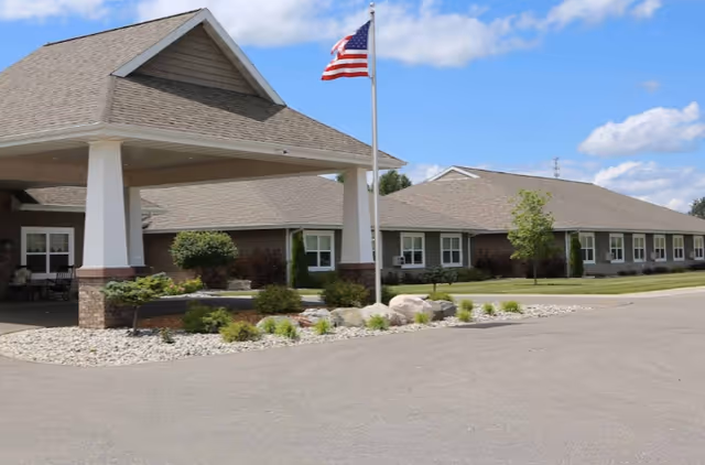 Exterior view of a single-story building with a covered entrance and an American flag on a flagpole in front. The building is surrounded by landscaping with bushes, small trees, and rocks under a partly cloudy sky.