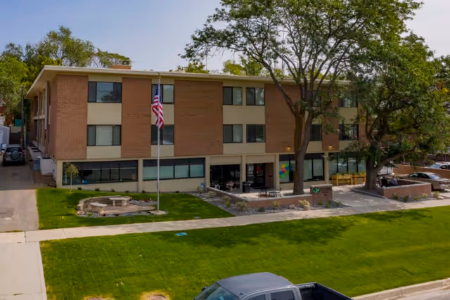 Exterior view of a three-story brick and beige building with multiple windows, an American flag on a flagpole in front, a small circular stone seating area, and large trees providing shade. There are parked cars visible on the right side and a green lawn in front.