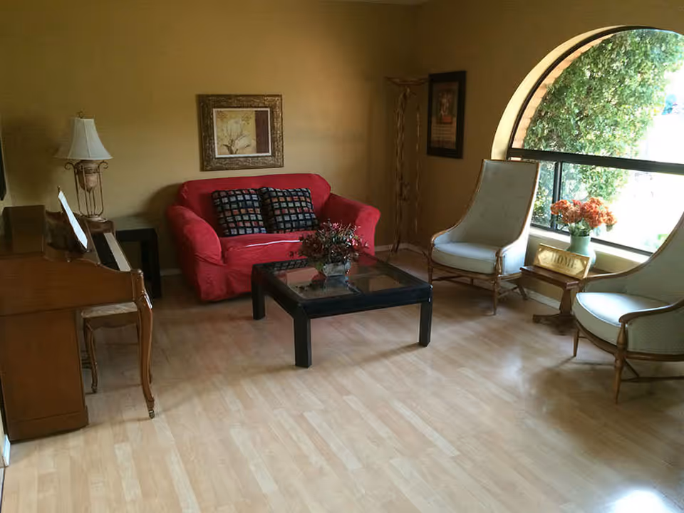 A cozy living room with a red loveseat adorned with two patterned cushions, a glass coffee table with a floral centerpiece, two white cushioned chairs, a wooden side table with a vase of orange flowers and a 'HOME' sign, a piano with a lamp on it, and a large arched window letting in natural light.