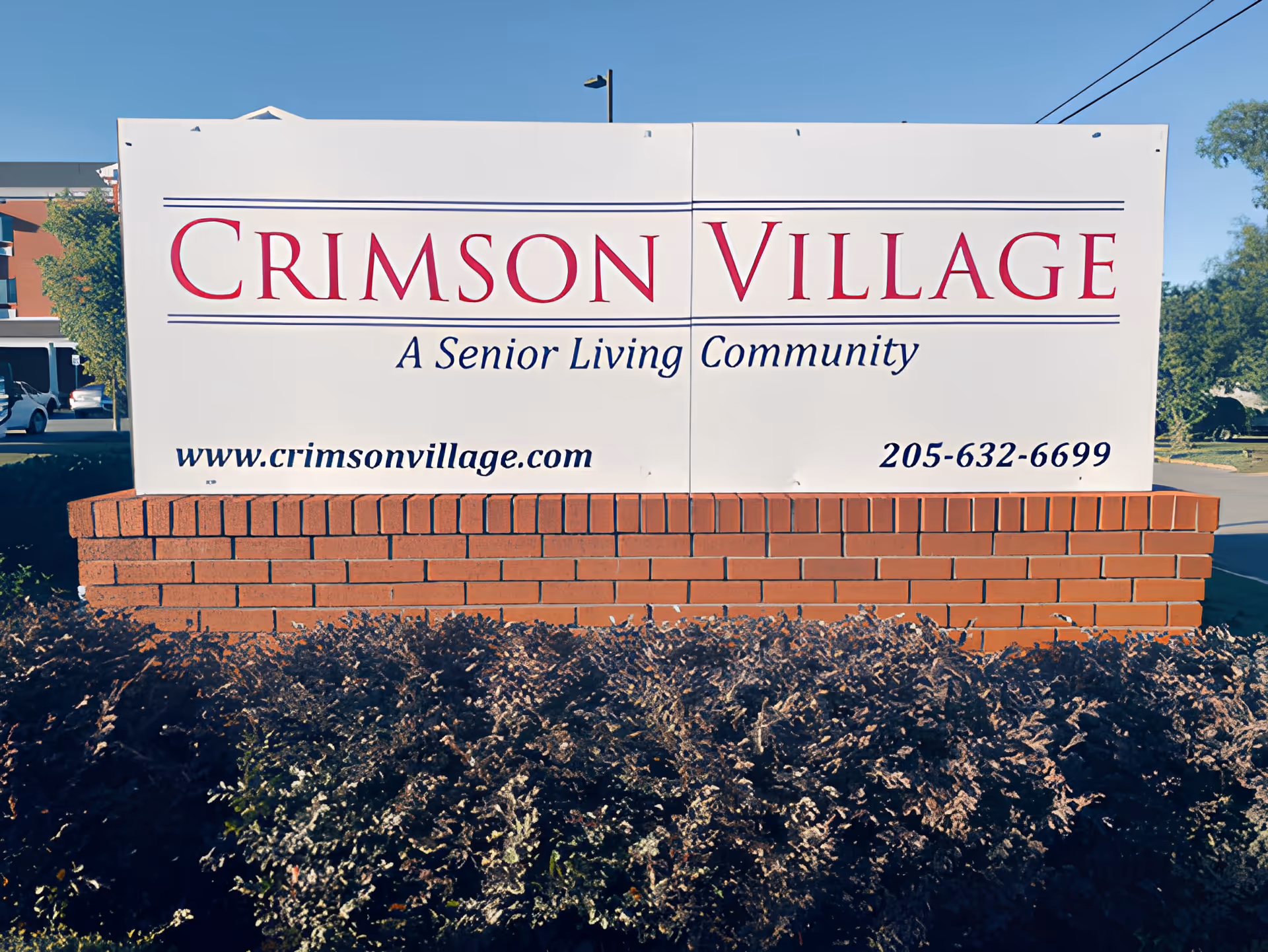 Outdoor sign for Crimson Village, a senior living community, mounted on a brick base with bushes in front and a clear blue sky in the background.