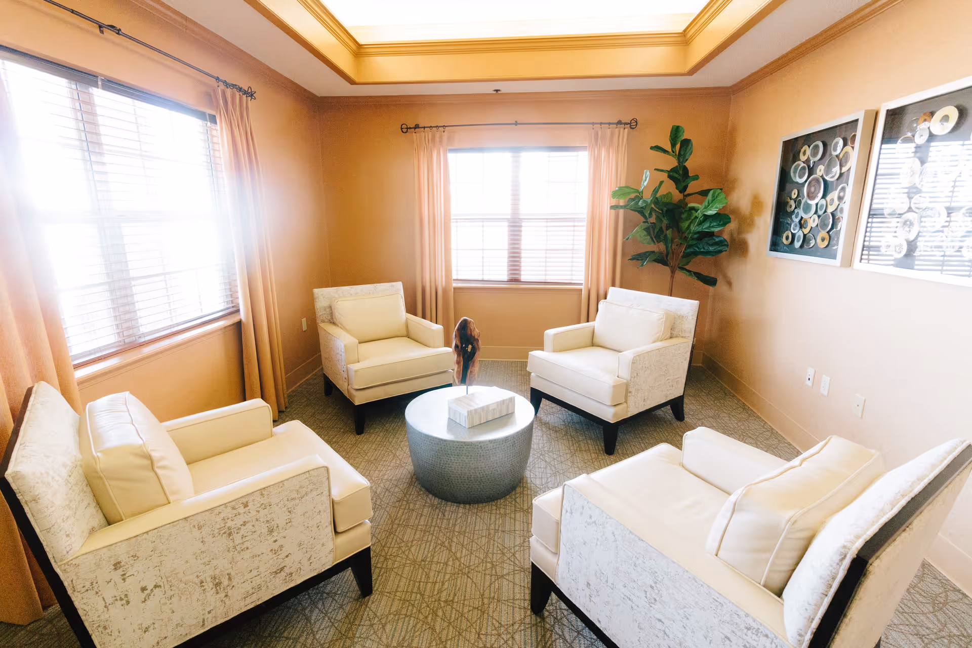 A bright and cozy sitting area with four cream-colored armchairs arranged around a round metallic coffee table. The room has beige walls, two windows with peach-colored curtains, a large green potted plant in the corner, and two framed decorative art pieces on the wall.