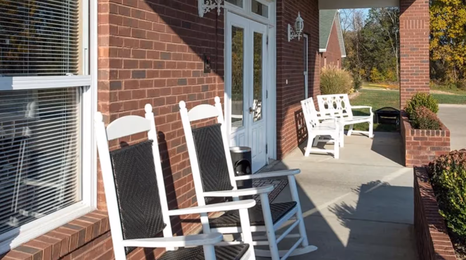 Covered brick front porch with white rocking chairs and benches outside the entrance of a senior living facility.
