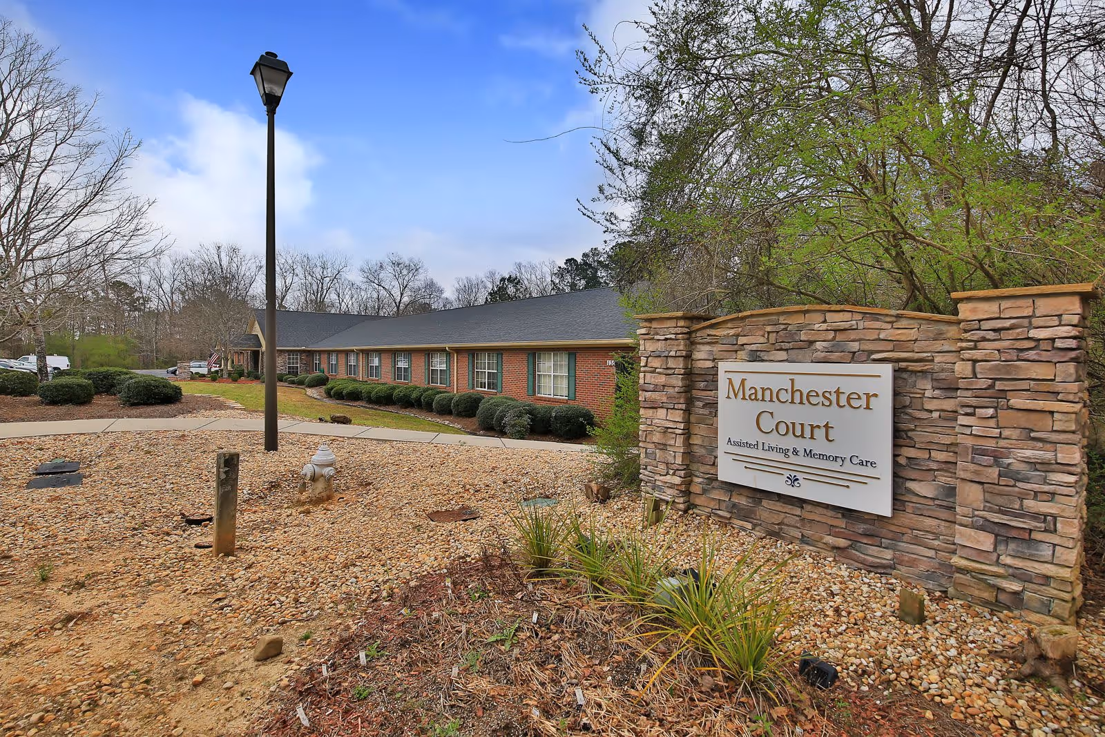 Exterior view of Manchester Court Assisted Living and Memory Care facility showing a brick building with multiple windows, a stone sign with the facility name, a lamppost, and surrounding landscaping with bushes and trees under a partly cloudy sky.