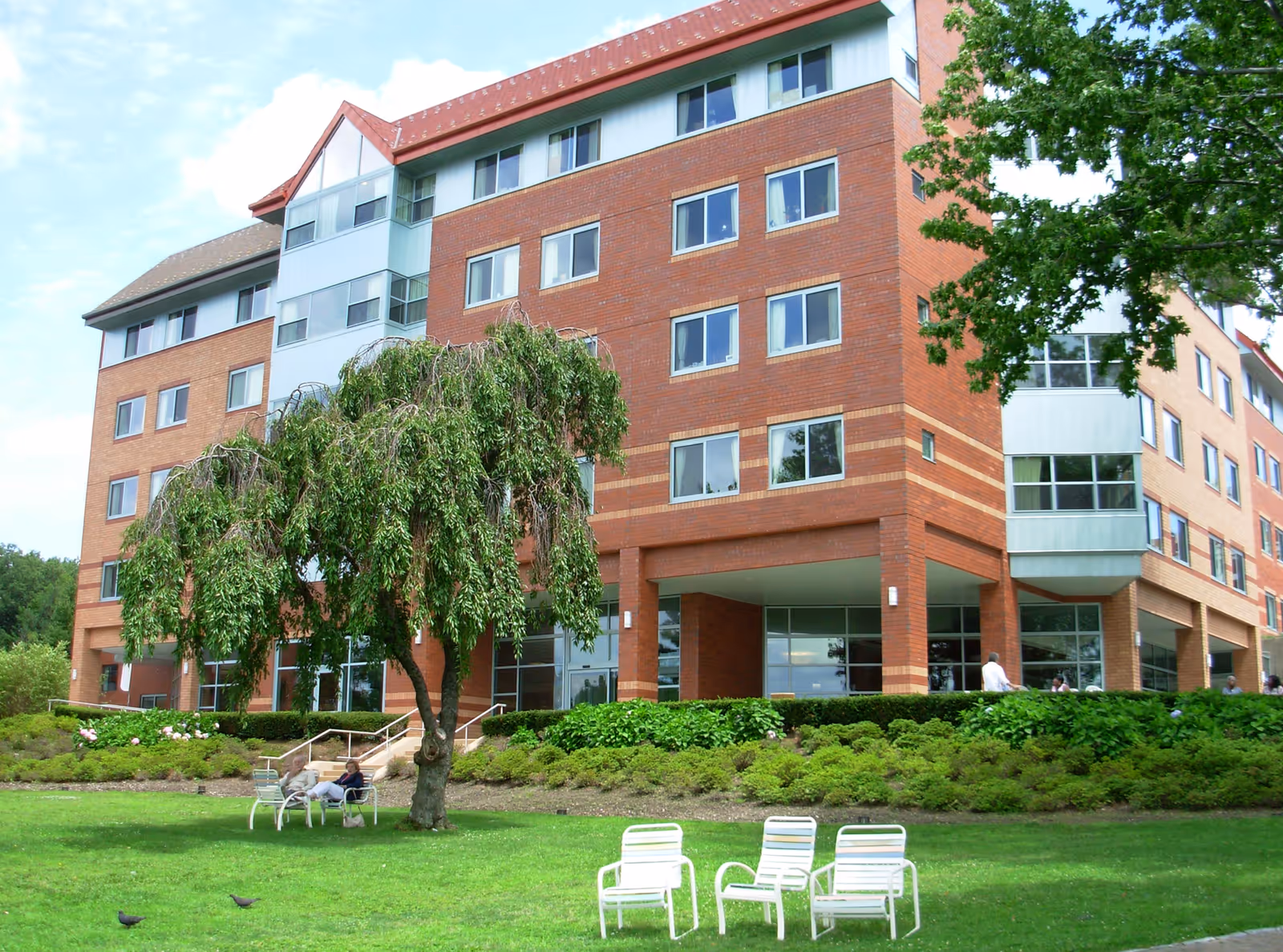 A multi-story brick building with large windows, surrounded by green shrubs and trees. In the foreground, there is a grassy lawn with white outdoor chairs and a small tree providing shade to two people sitting on a bench. The sky is partly cloudy.