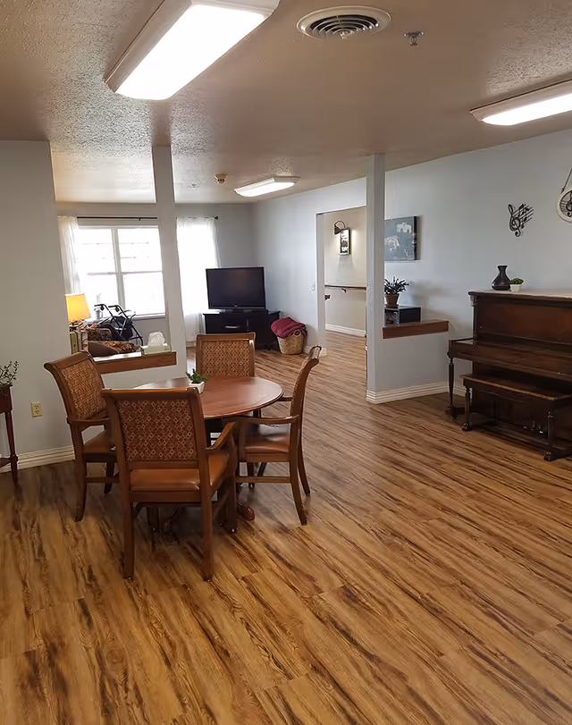 Bright communal room with a round wooden dining table and chairs, a TV, piano and wood-look flooring.
