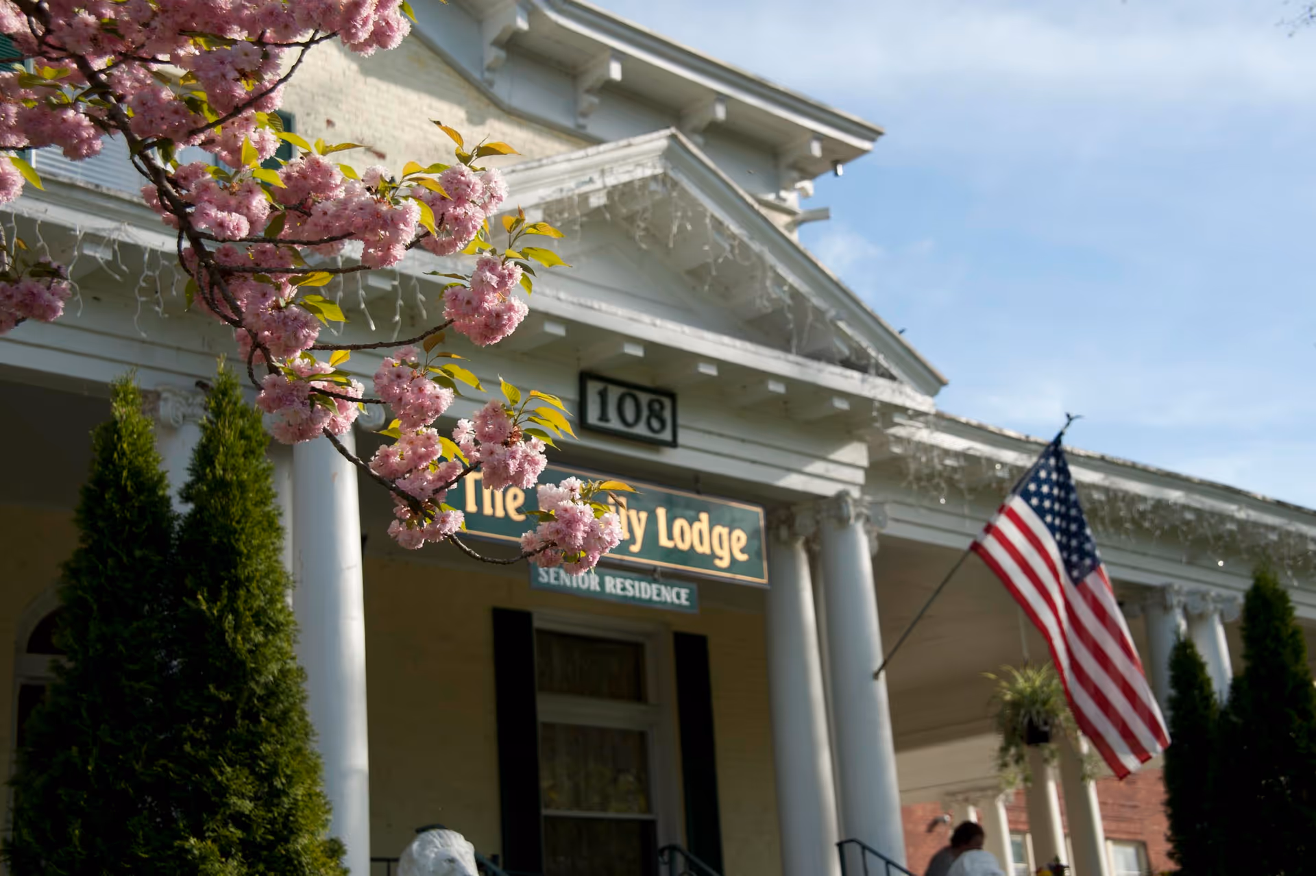 Front view of Ivy Lodge Assisted Living building with pink blossoms and a flag