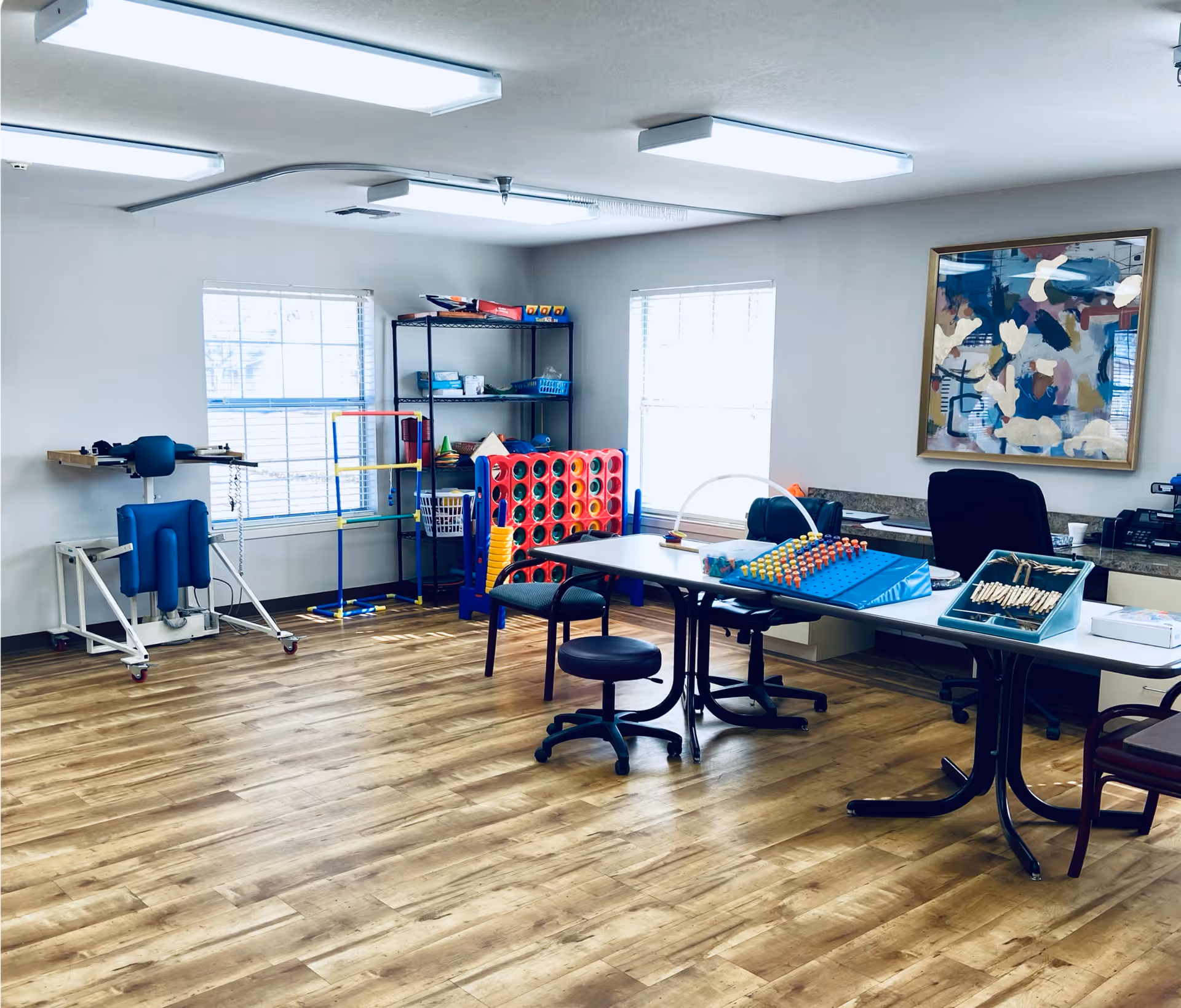 A bright room with wooden flooring featuring a long table with chairs and various activity and therapy equipment, including a large Connect Four game, pegboards, and other cognitive and physical therapy tools. There are two windows letting in natural light, a metal shelving unit with supplies, and an abstract painting on the wall.