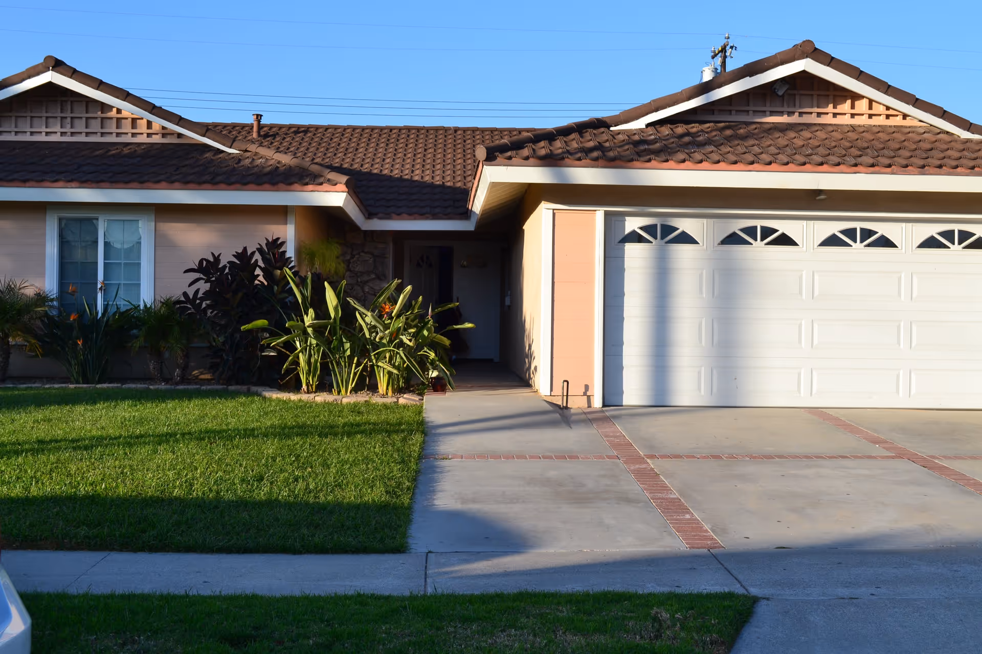 Front exterior of a single-story house with a two-car garage, driveway, lawn, and plants.