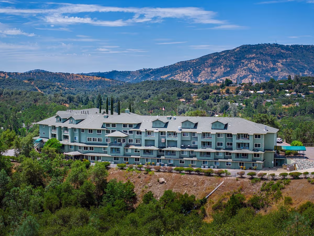 A light-green senior living building sits along a hillside with mountains in the background.