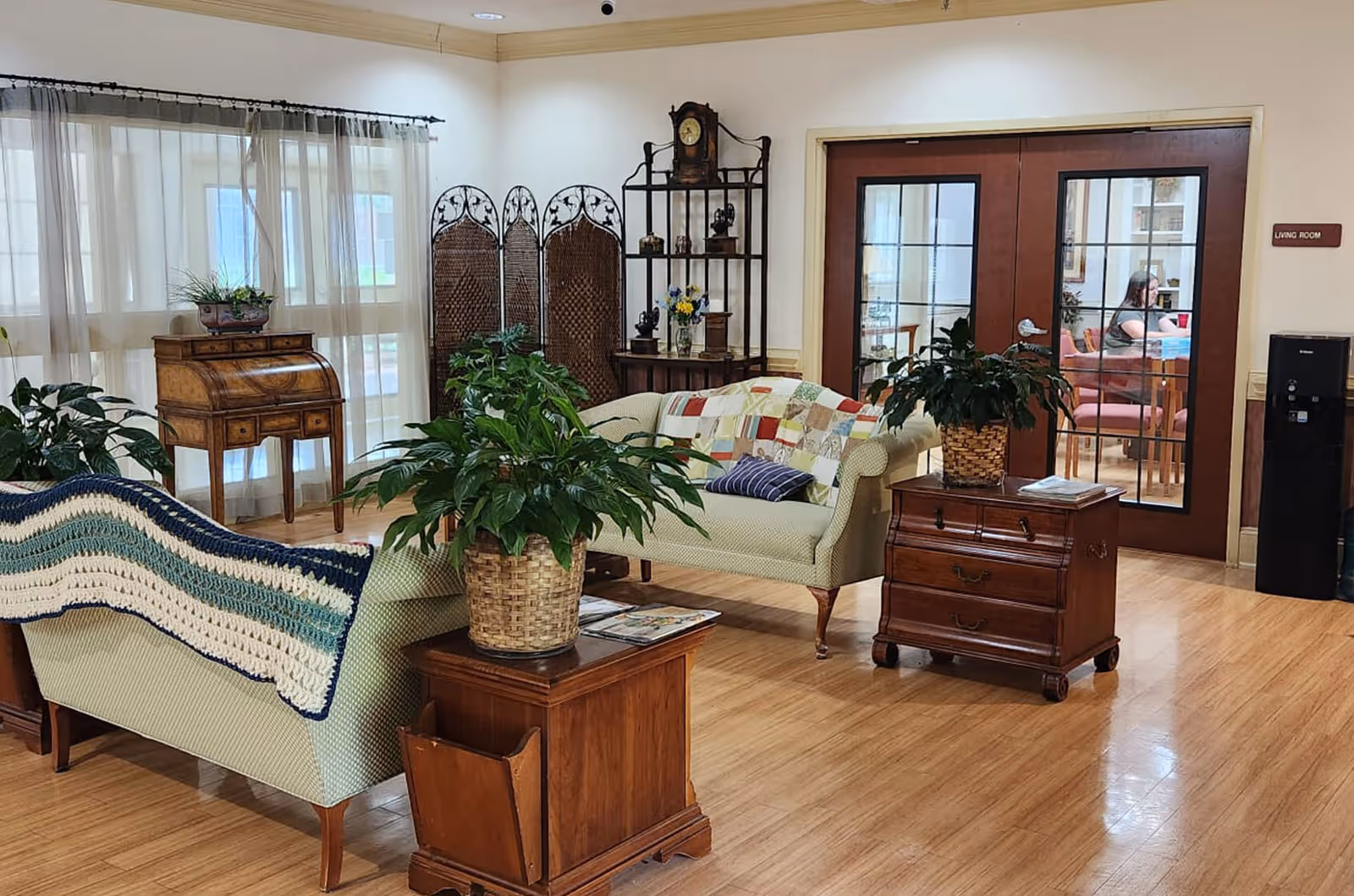 A cozy living room area with two light green upholstered sofas, one draped with a blue and white crocheted blanket and the other with a colorful patchwork quilt. There are two wooden side tables with potted plants, a wooden roll-top desk near sheer curtained windows, a decorative folding screen, and a tall wooden shelving unit with a clock and small decorative items. In the background, double glass doors lead to another room where a person is seated at a table. The floor is wooden, and a water cooler is visible near the doors.
