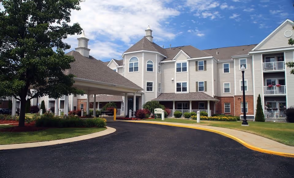 Exterior view of NorthPointe Woods senior living facility showing a multi-story building with beige siding and brick accents, a covered entrance driveway, landscaped greenery, and a partly cloudy sky.