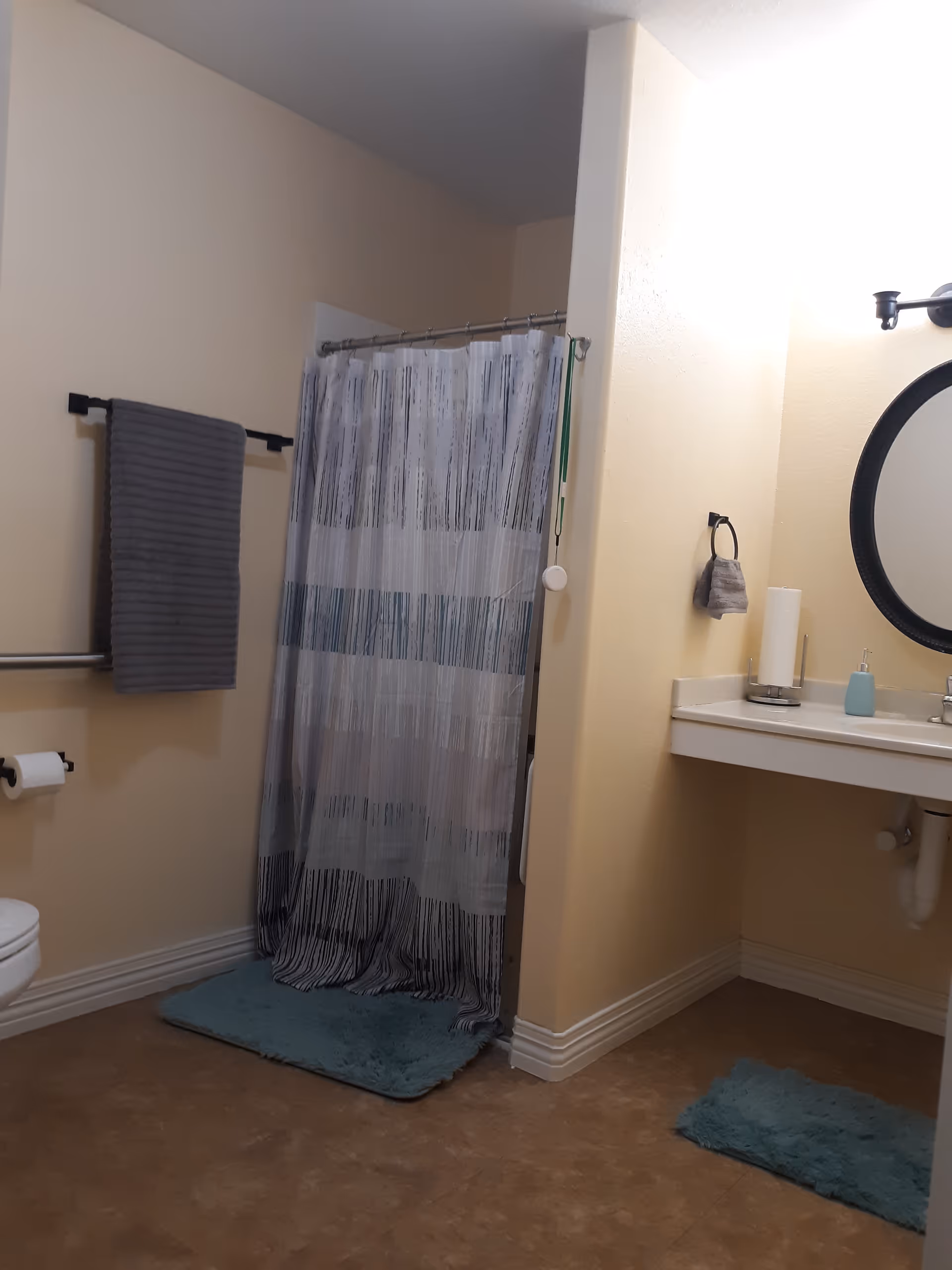 Bathroom in a senior living facility with shower curtain, sink, mirror, towels, and blue mats.