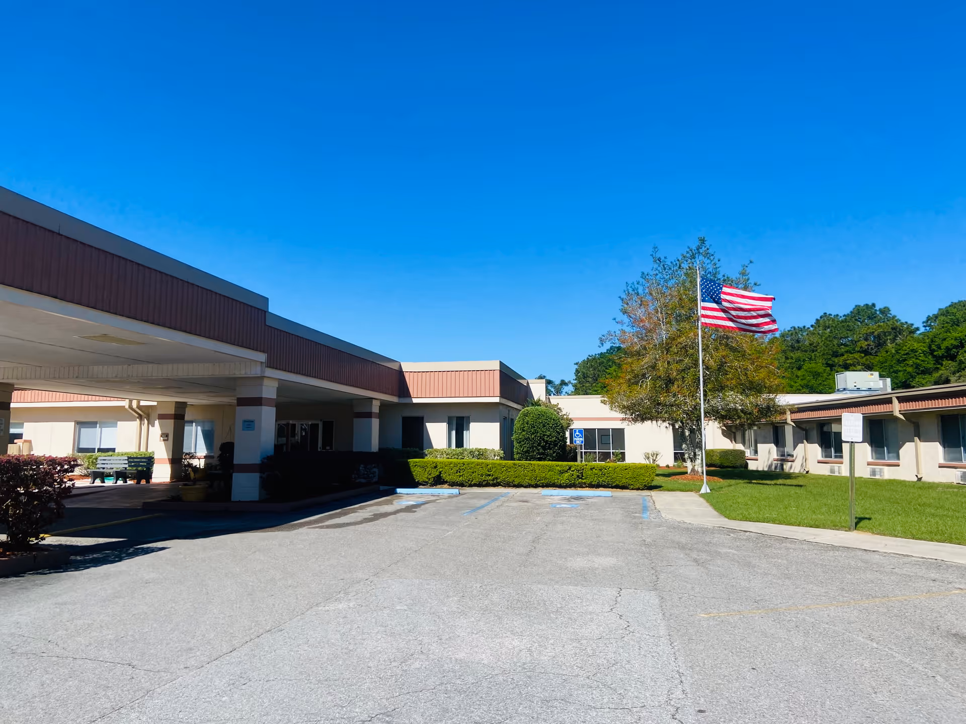 Exterior view of Tri-County Nursing Home building with a covered entrance, parking spaces including handicapped spots, an American flag on a flagpole, green bushes, and trees under a clear blue sky.