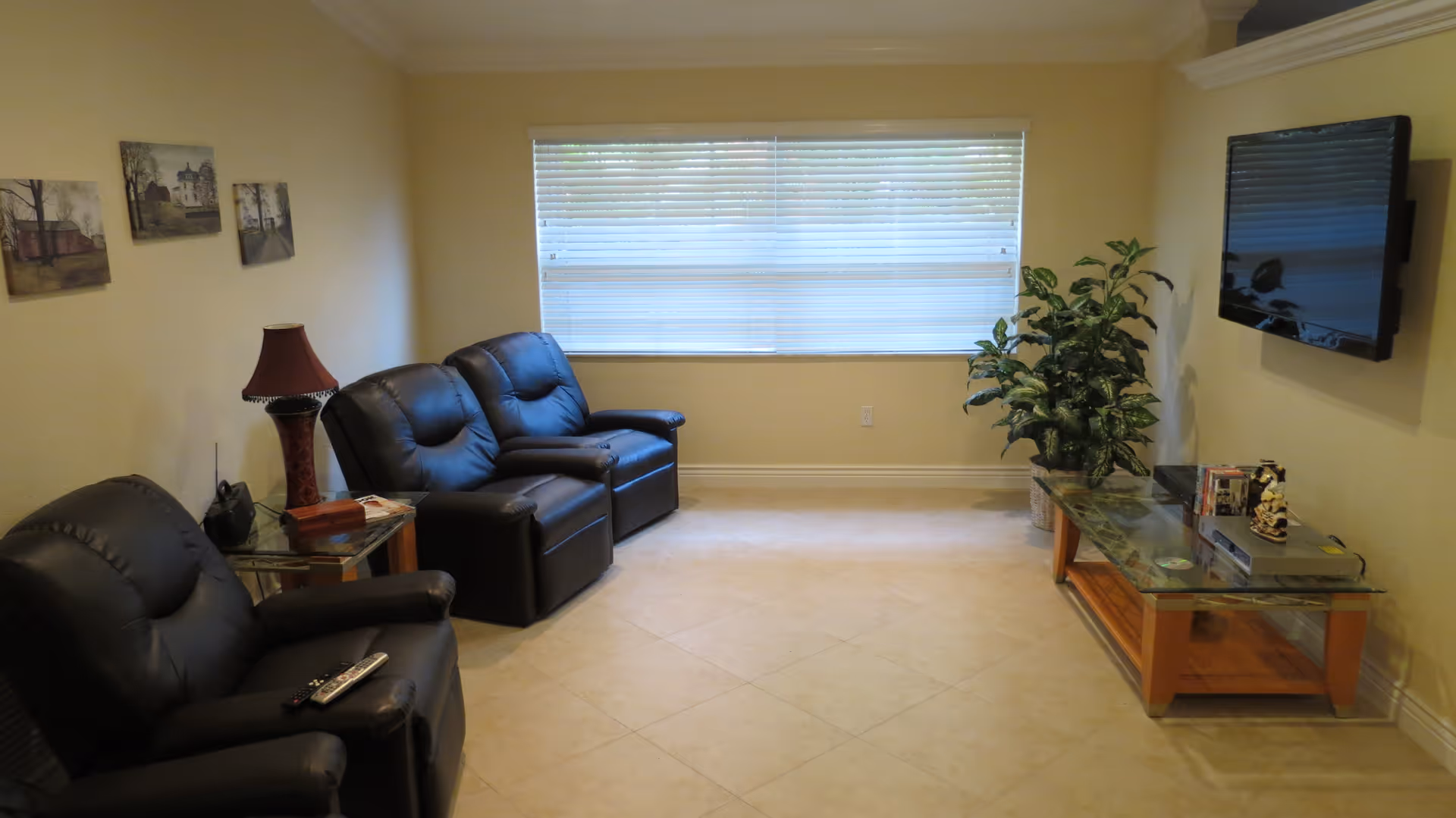 A cozy living room with three black leather recliner chairs arranged along the walls. A glass-top wooden coffee table holds decorative items and is positioned under a wall-mounted flat-screen TV. A large window with blinds lets in natural light, and a green potted plant adds a touch of nature to the room. The walls are painted a light beige color and have three framed pictures hanging on one side.