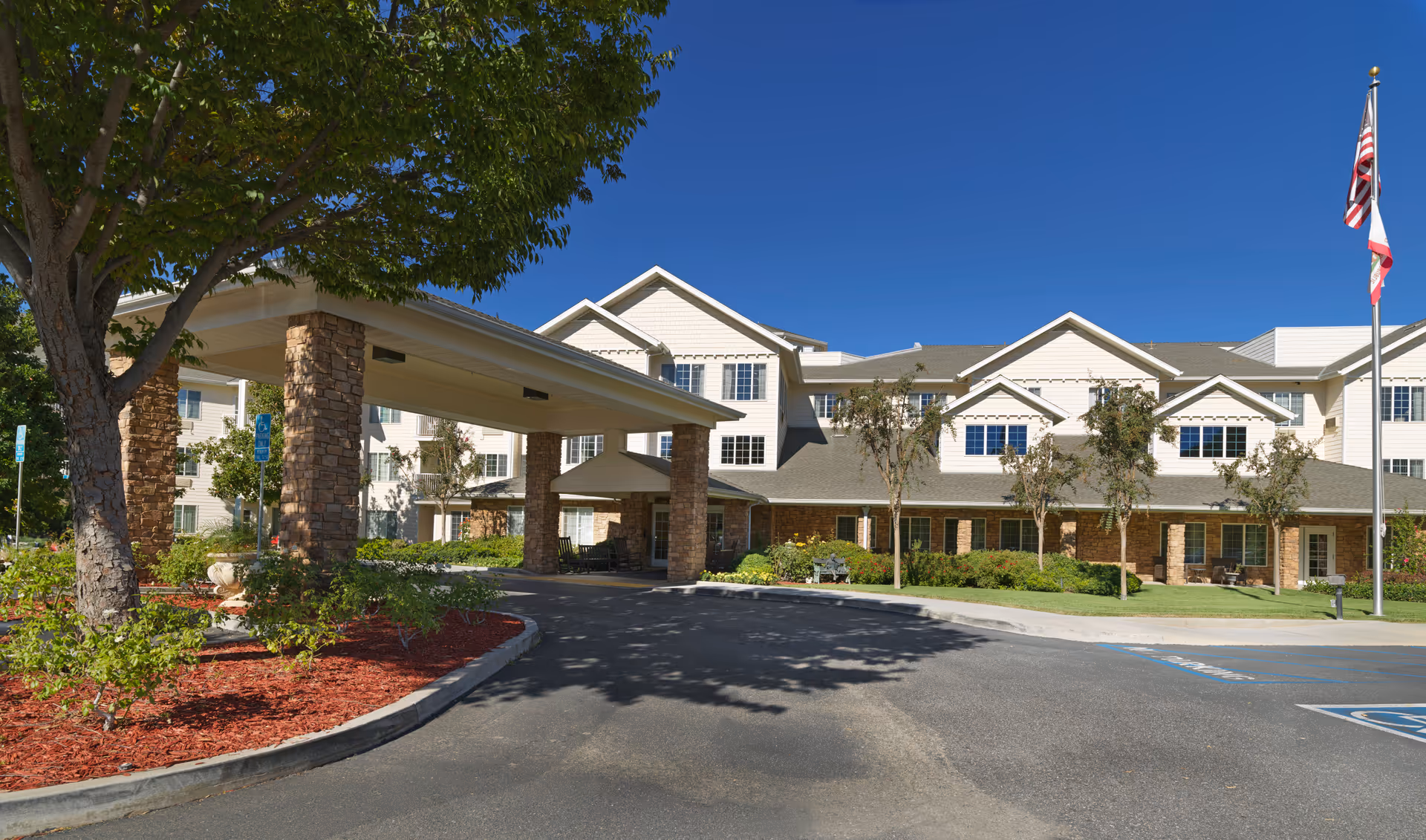 Front view of a multi-story senior living building with a covered entrance porte-cochere, landscaped grounds, and flags.