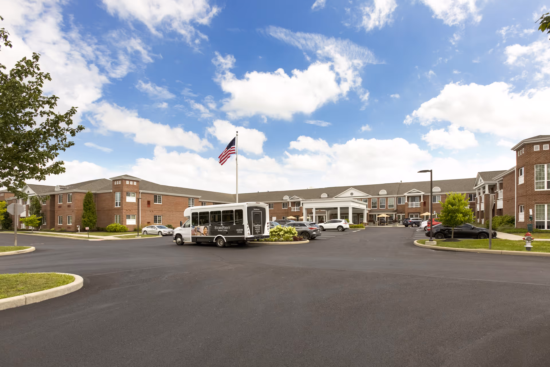 Exterior view of StoryPoint Gahanna North senior living facility showing a large brick building with multiple windows, a covered entrance, several parked cars, a shuttle bus, an American flag on a flagpole, and a partly cloudy sky.