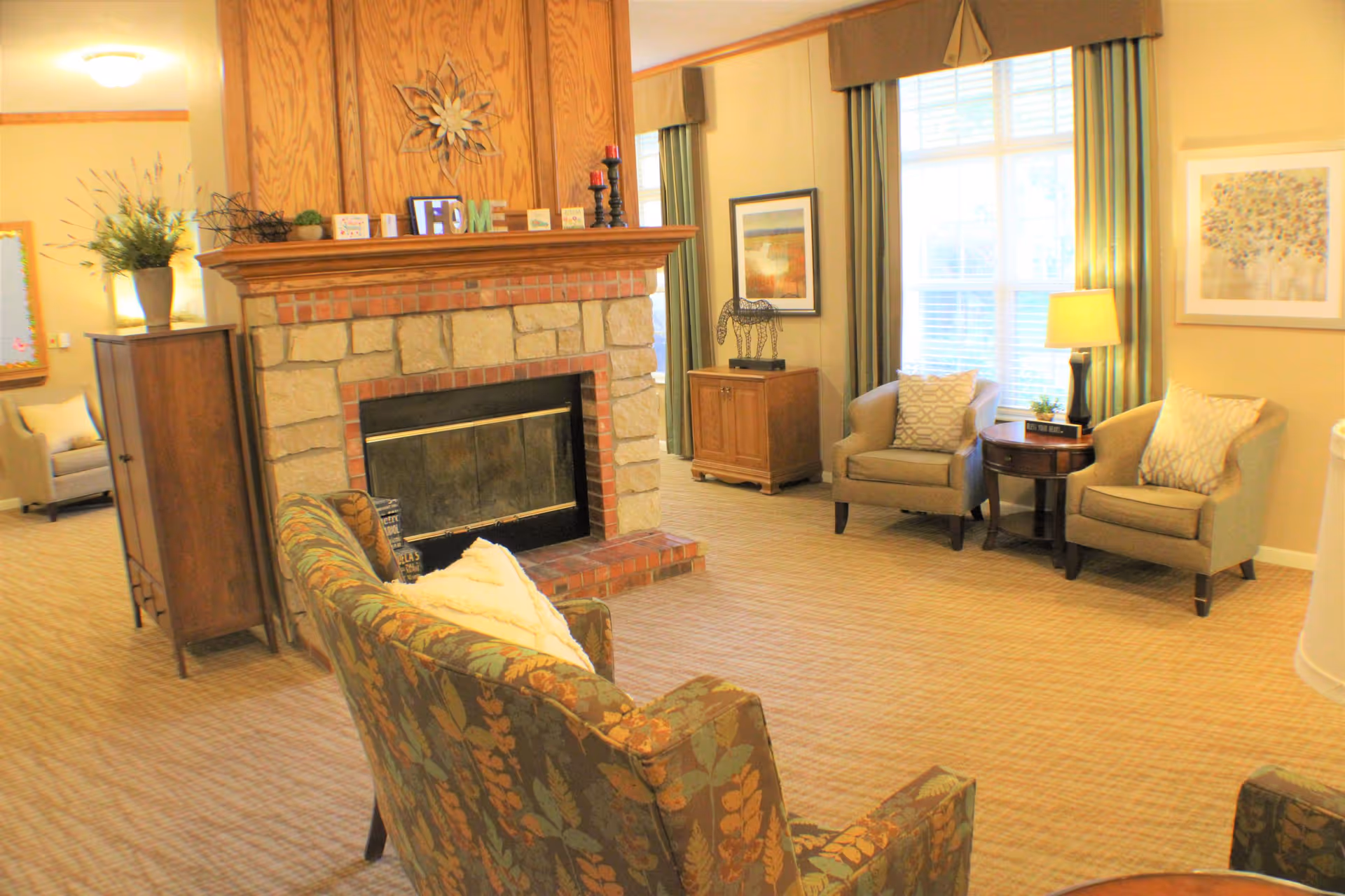 Comfortable common living room with a stone fireplace, upholstered chairs and sofas, side tables and large windows.