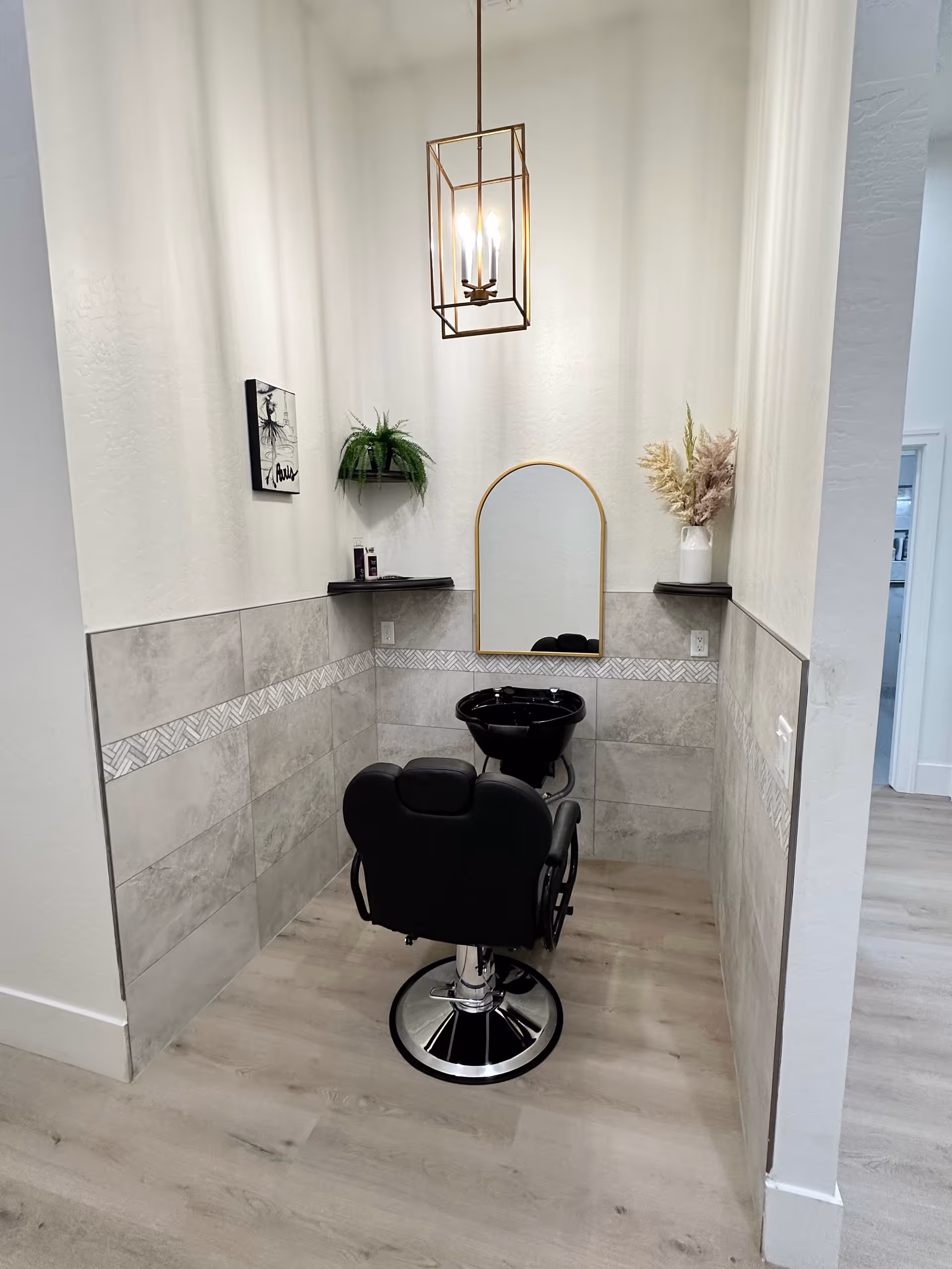 A small salon hair-washing station with a black styling chair, shampoo sink, arched mirror, and pendant light in a tiled alcove.