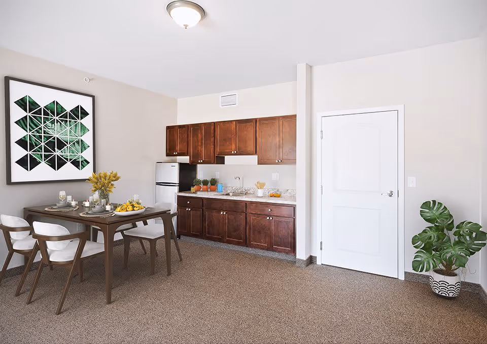 A small kitchen and dining area with dark wood cabinets, a white refrigerator, and a countertop with various items including plants and kitchenware. A wooden dining table with four white cushioned chairs is set with plates, glasses, and a centerpiece of yellow flowers and fruit. A large framed geometric artwork hangs on the wall above the dining table. A white door and a potted plant are visible on the right side of the room.