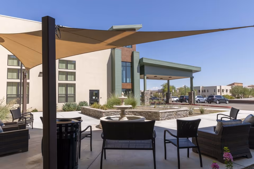 Outdoor seating area with shade sails and a fountain in front of the entrance of a modern senior living building.
