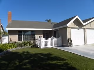 Single-story beige senior living unit with a white picket railing, front porch, and green lawn.
