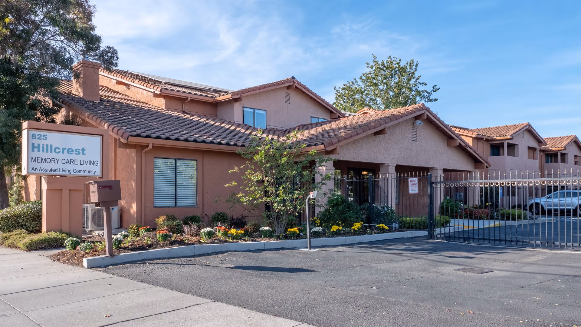 Exterior view of a single-story assisted living community building with a tiled roof, surrounded by landscaping with flowers and bushes. A sign in front reads '825 Hillcrest Memory Care Living An Assisted Living Community'. There is a gated entrance to the parking area on the right side of the building under a clear blue sky.
