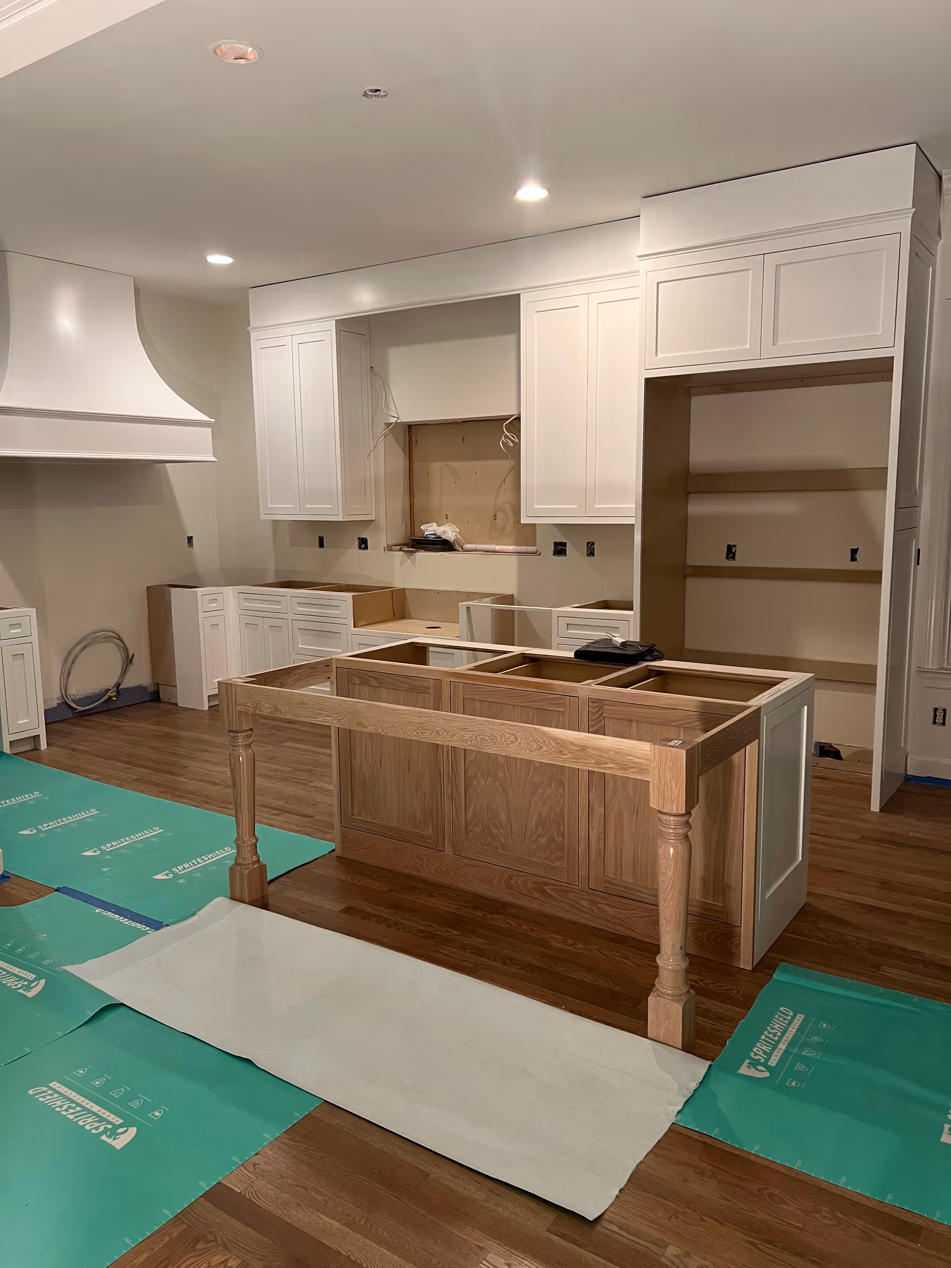 Under-construction kitchen with white cabinets, an island frame, and protective green floor coverings.