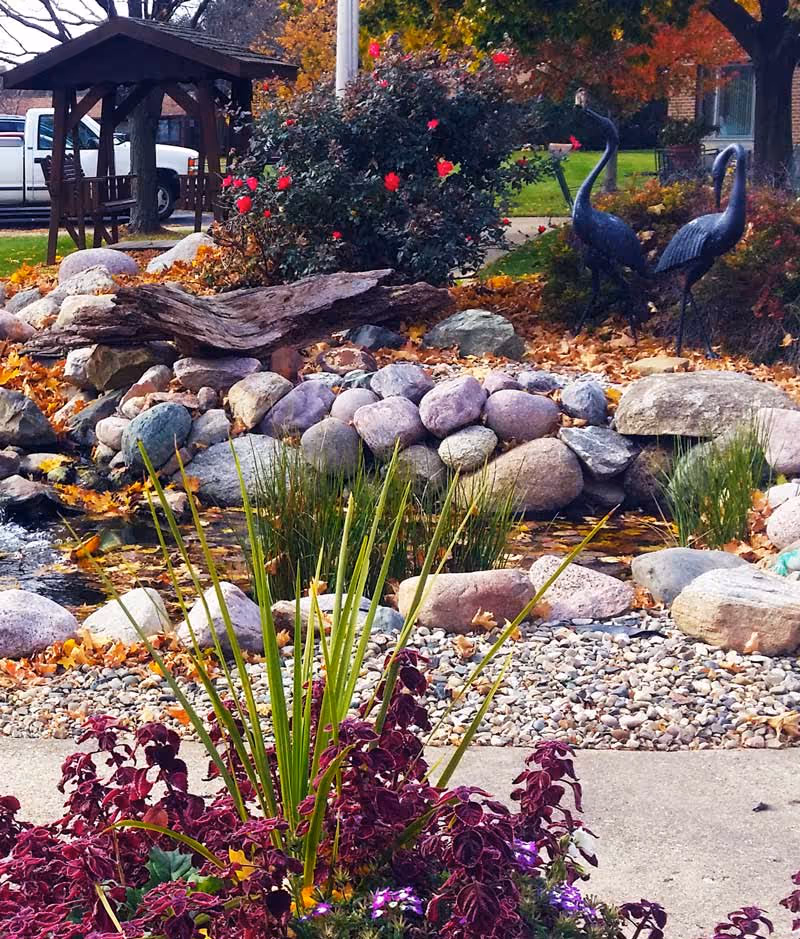 A landscaped outdoor garden area featuring a small rock pond surrounded by various stones and plants. There are two metal crane sculptures near the pond, colorful flowers and bushes, and a wooden gazebo with a bench in the background. Fallen autumn leaves are scattered around the scene.