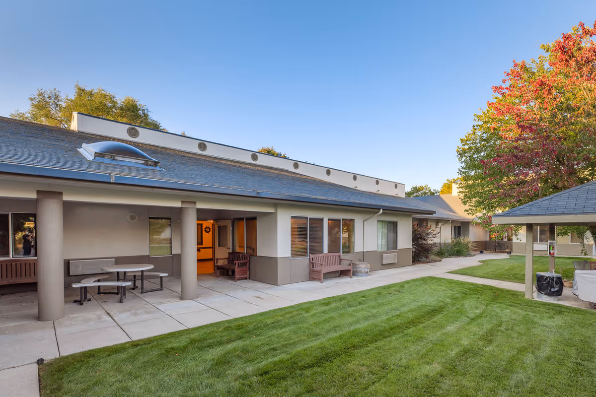 Outdoor courtyard showing a grassy lawn, covered patio with benches and tables, and the exterior of a one-story senior living building.