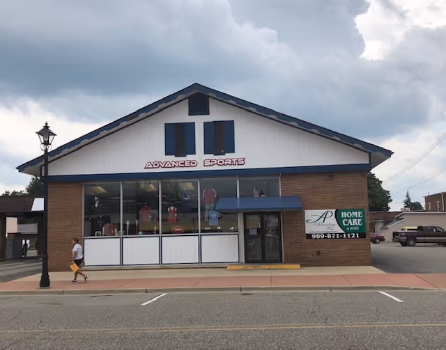 Brick storefront with large display windows and Advanced Sports sign; person walking on sidewalk.