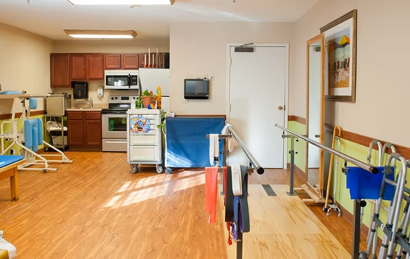 A rehabilitation room with parallel bars for walking exercises, resistance bands hanging on the bars, a large mirror on the wall, and a kitchen area with wooden cabinets, a microwave, stove, and sink in the background. The floor is wooden, and there is a cart with supplies near the kitchen.