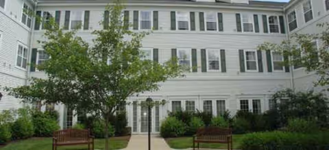 White multi-story senior living building with trees, benches, and a central walkway.