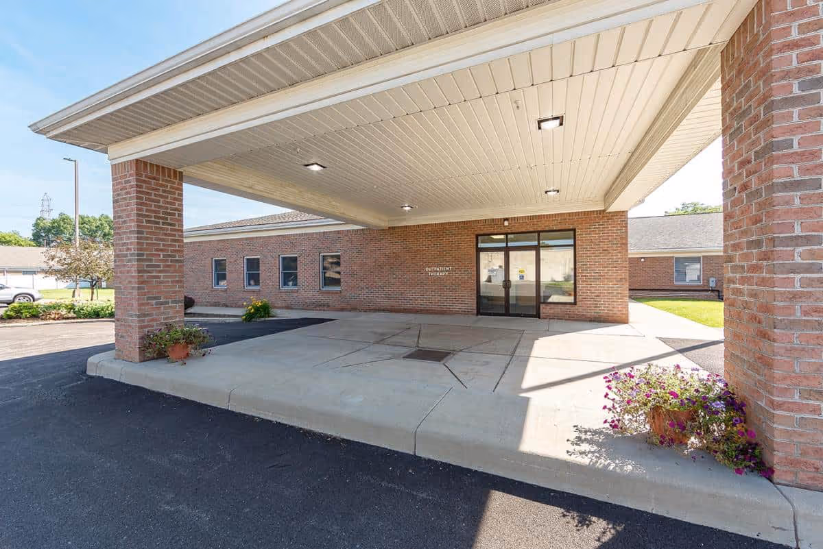 Covered entrance area of a brick building with a concrete driveway and two brick pillars supporting the roof. There are potted plants with flowers near the pillars and a glass door entrance labeled 'Outpatient Therapy'.