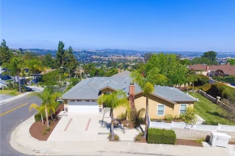 Aerial view of a single-story suburban house with a driveway, palm trees, and a distant cityscape.