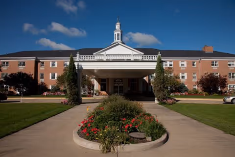 Brick senior living facility with front entrance, manicured lawn, and central circular flower bed.
