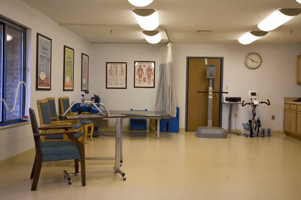 A therapy room with chairs, a wheeled table, exercise equipment, and framed medical posters.