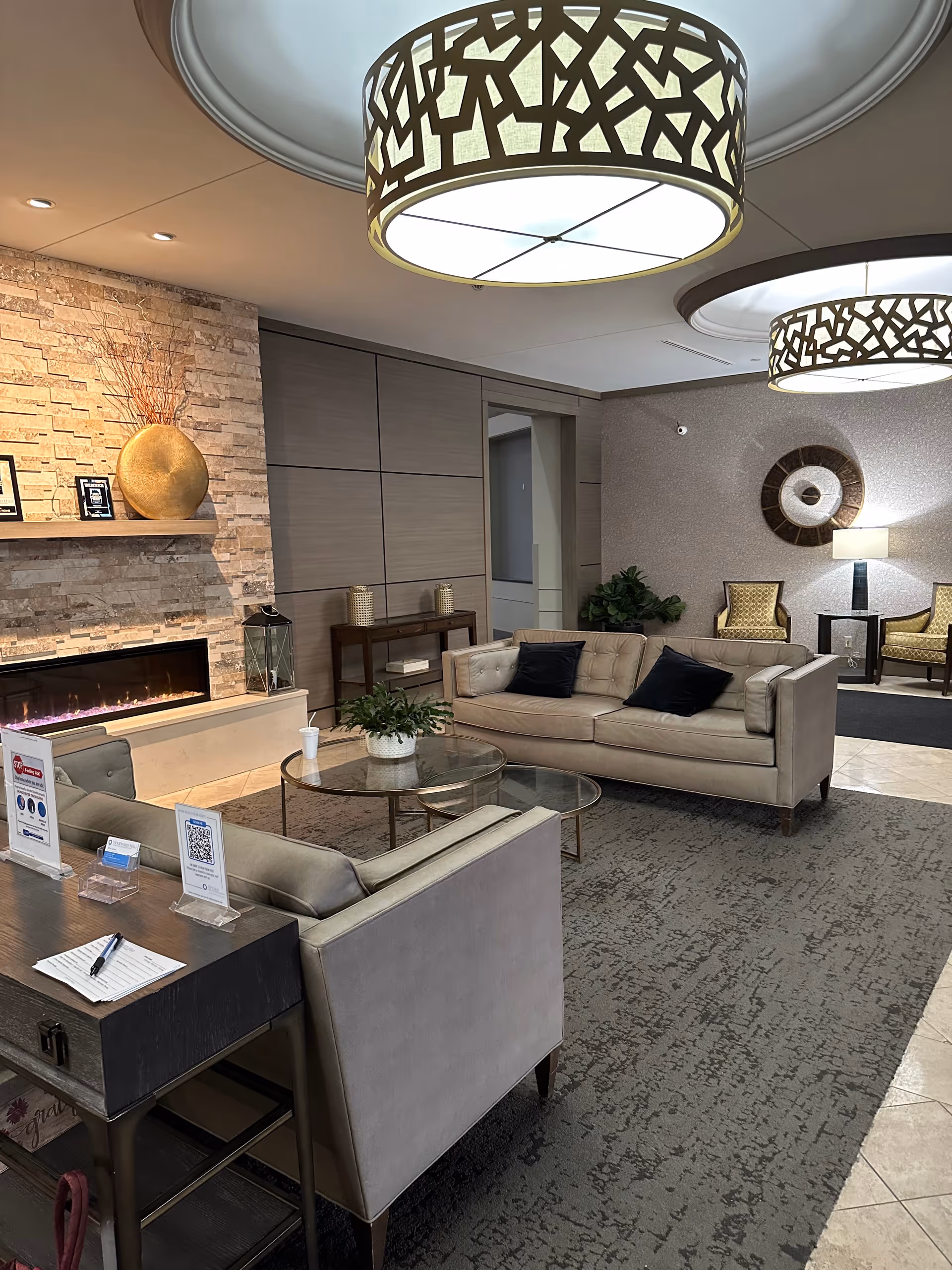 Modern senior living lobby with beige seating, glass tables, a fireplace, and decorative lighting.