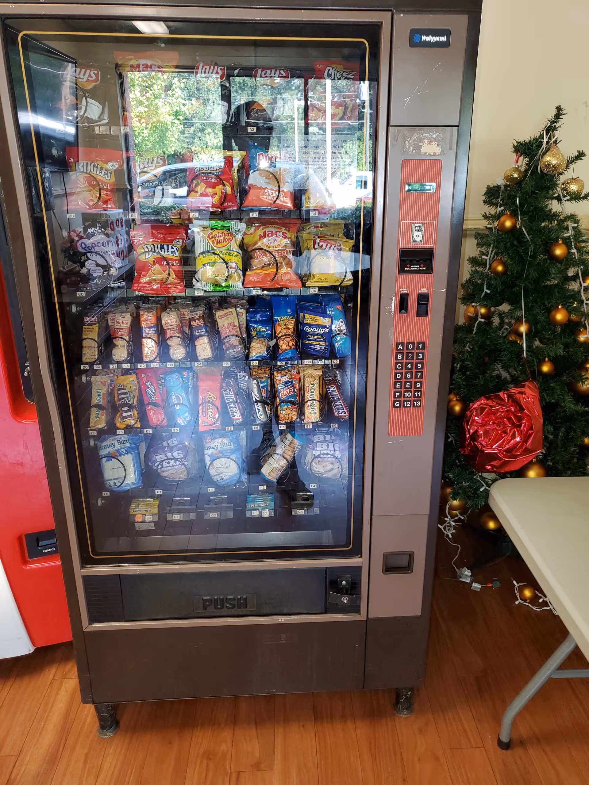 Snack vending machine filled with chips inside Abernathy Towers near a Christmas tree.