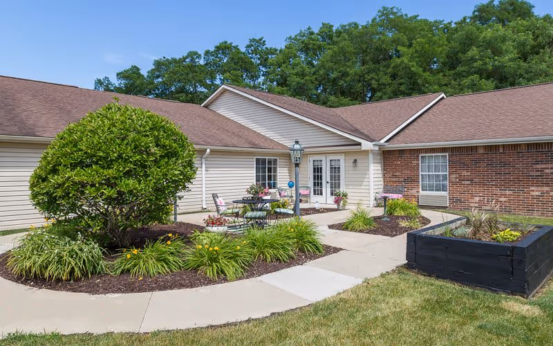 Sunny courtyard with patio tables and chairs, landscaped planting beds, and the single-story facility exterior with French doors.