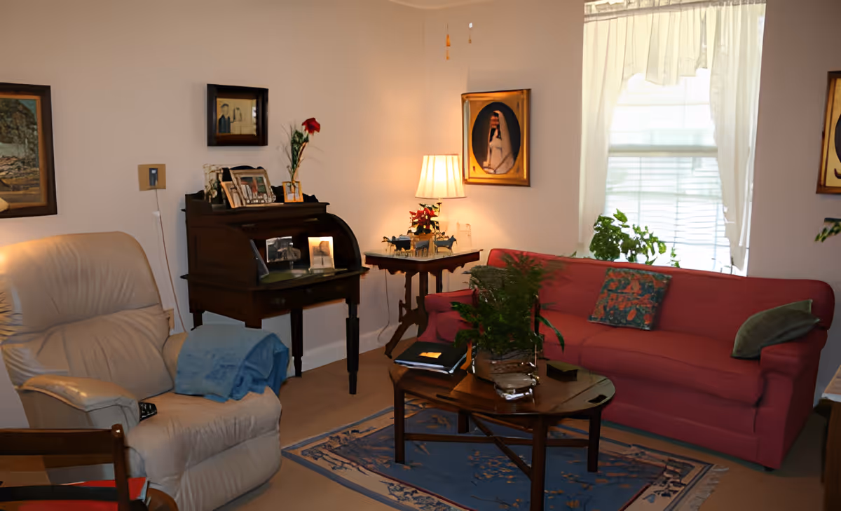 A cozy living room with a red sofa adorned with two cushions, a beige recliner chair with a blue blanket draped over one arm, a wooden coffee table with a potted plant and books, a small wooden side table with a lamp and decorative items, and framed artwork on the walls. A window with sheer white curtains allows natural light into the room.