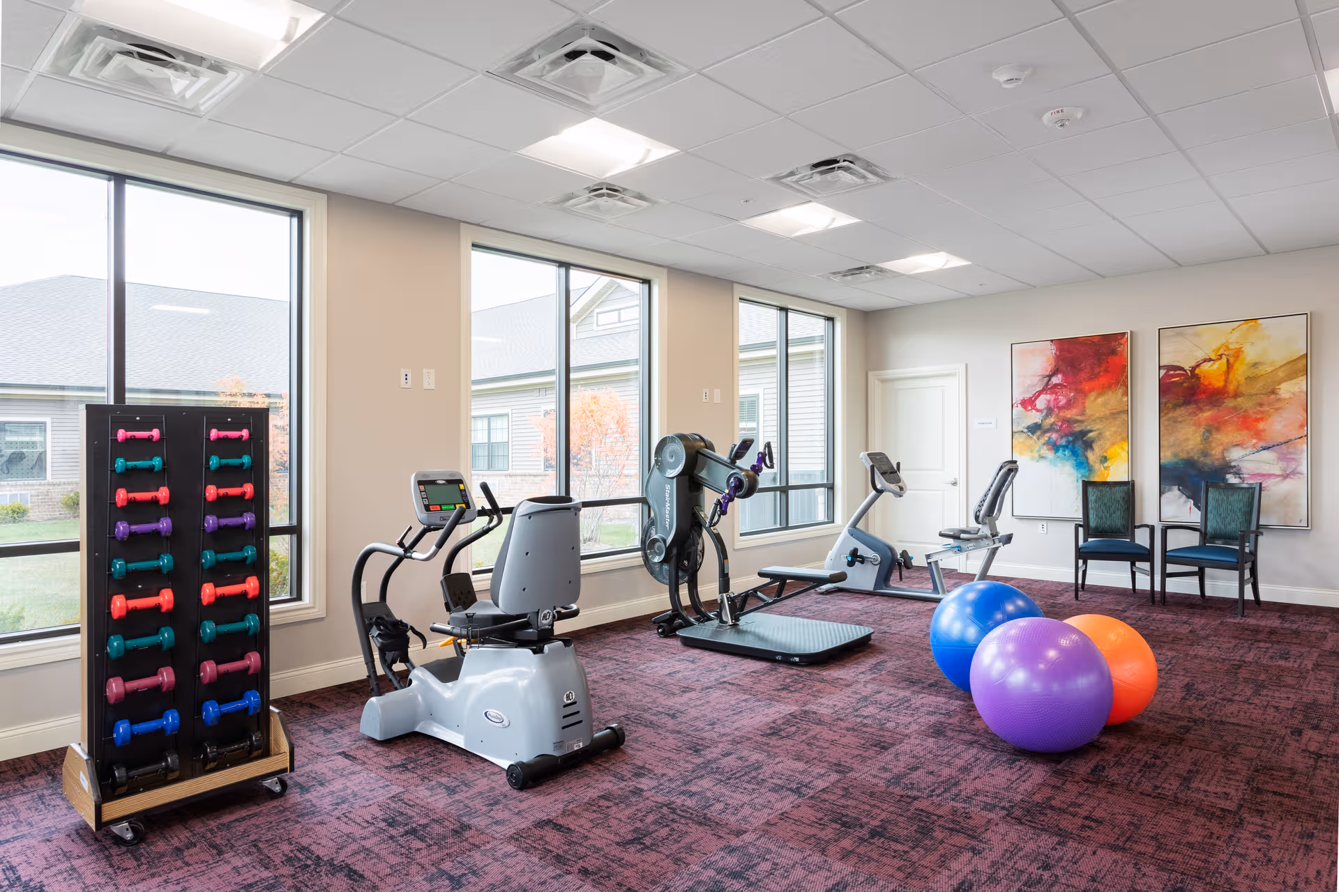 Senior living fitness room with treadmill, stationary bike, weights, exercise balls, and colorful abstract art.