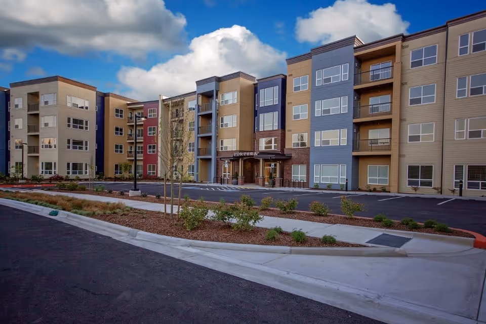 Exterior view of a modern multi-story senior living facility named Portera at the Grove, featuring a landscaped entrance with small trees and shrubs, a parking area, and a partly cloudy sky.