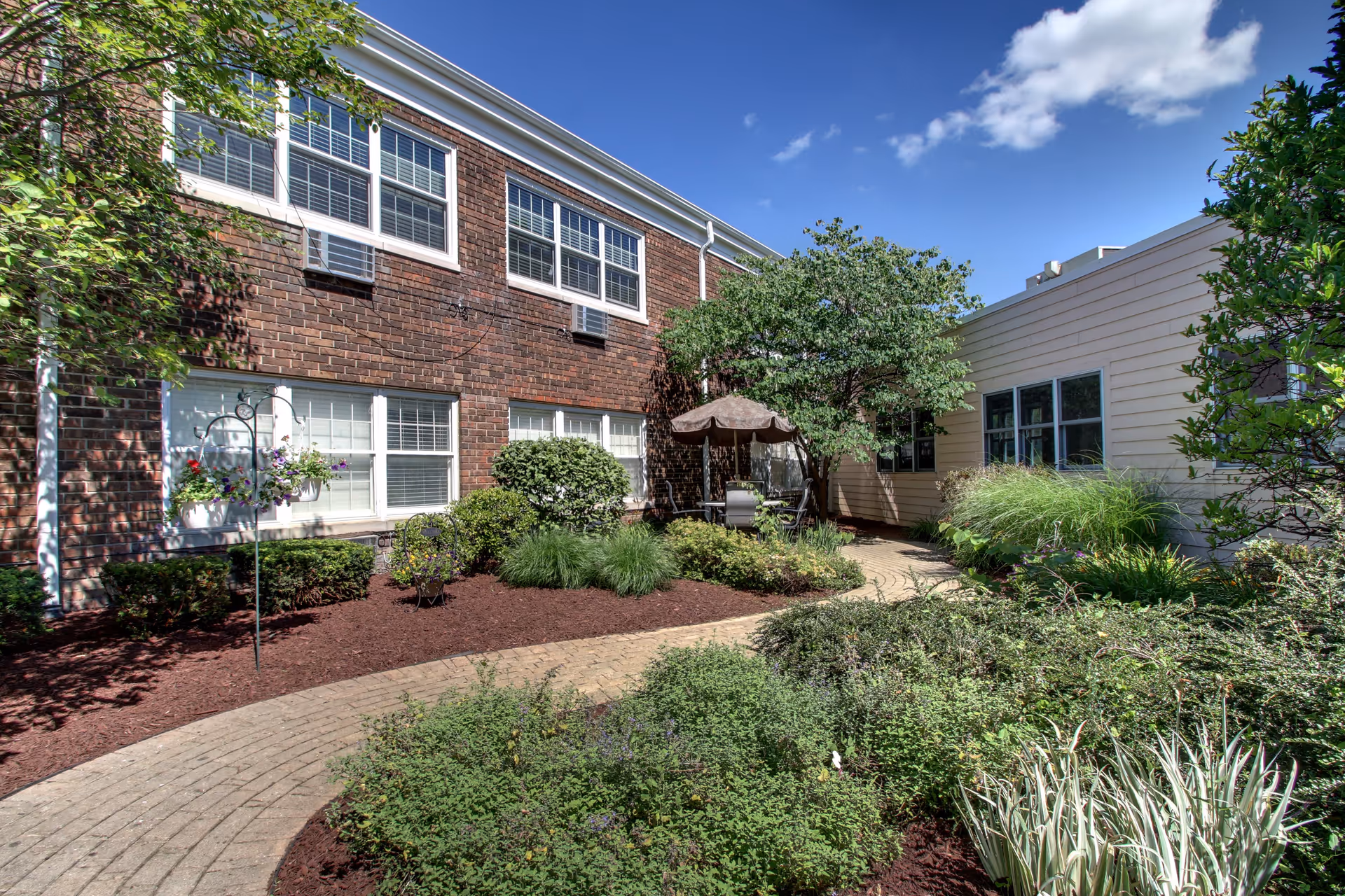 Outdoor garden area at BRIA of Geneva featuring a curved brick pathway surrounded by various green shrubs and plants. There is a brick building on the left with multiple windows and air conditioning units, and a light-colored building on the right. A patio table with chairs and a large umbrella is visible near the center under a tree. The sky is clear with a few clouds.