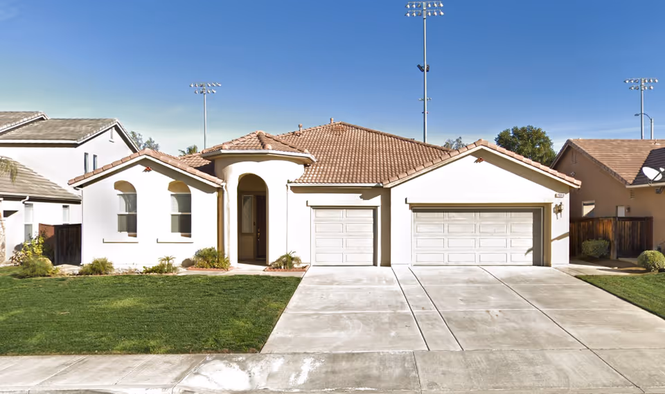 Front exterior view of a single-story house with a tiled roof, three garage doors, a small lawn, and a concrete driveway under a clear blue sky.