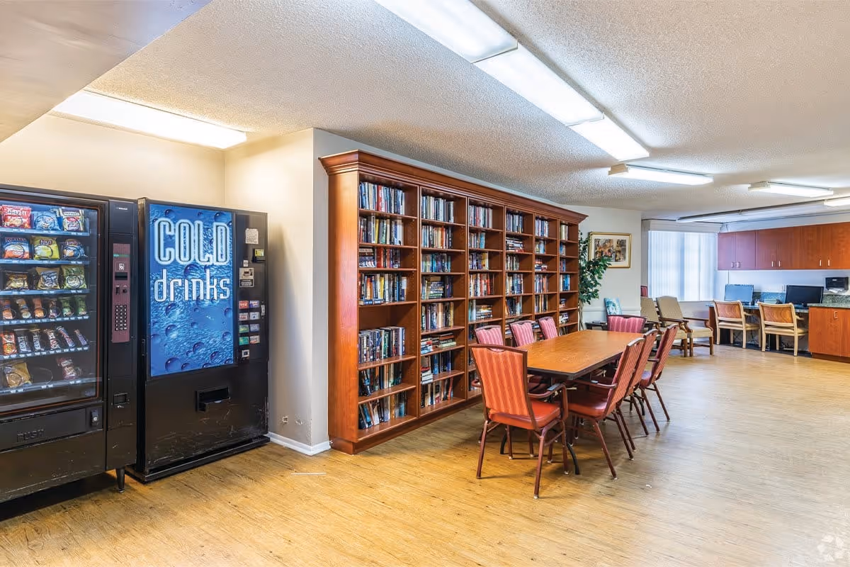 A senior living community common area featuring a vending machine with snacks and cold drinks, a large wooden bookshelf filled with books, a table with several red cushioned chairs, and a computer station with chairs along the back wall. The room has wood flooring and fluorescent ceiling lights.