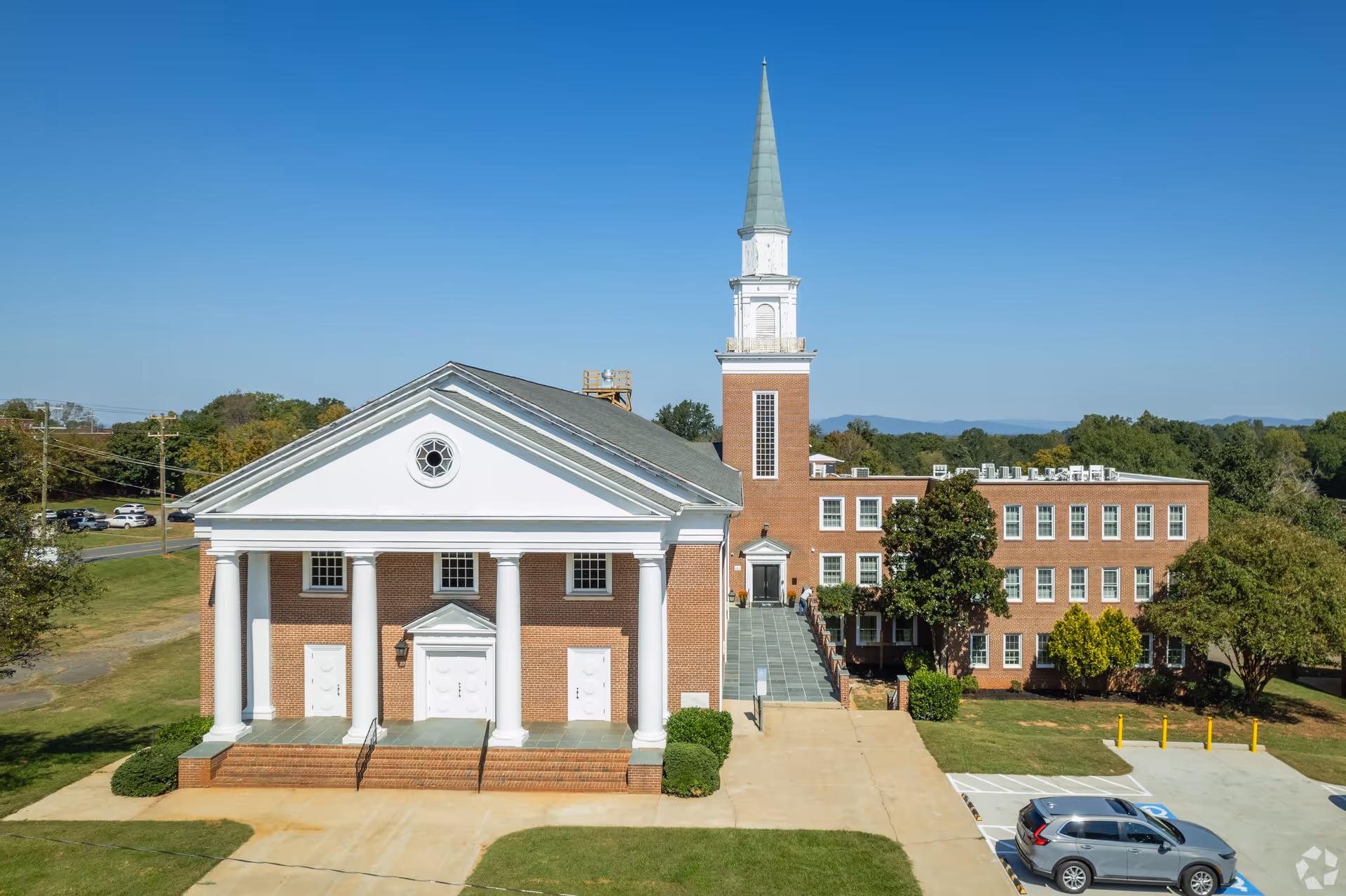 Exterior view of a large brick building with white columns and a tall steeple under a clear blue sky. There is a parking area with a car in front and green trees surrounding the building.