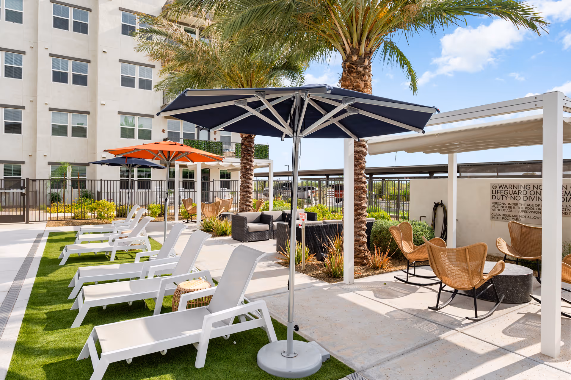 Outdoor lounge area with white lounge chairs, umbrellas, palm trees, and a modern building.