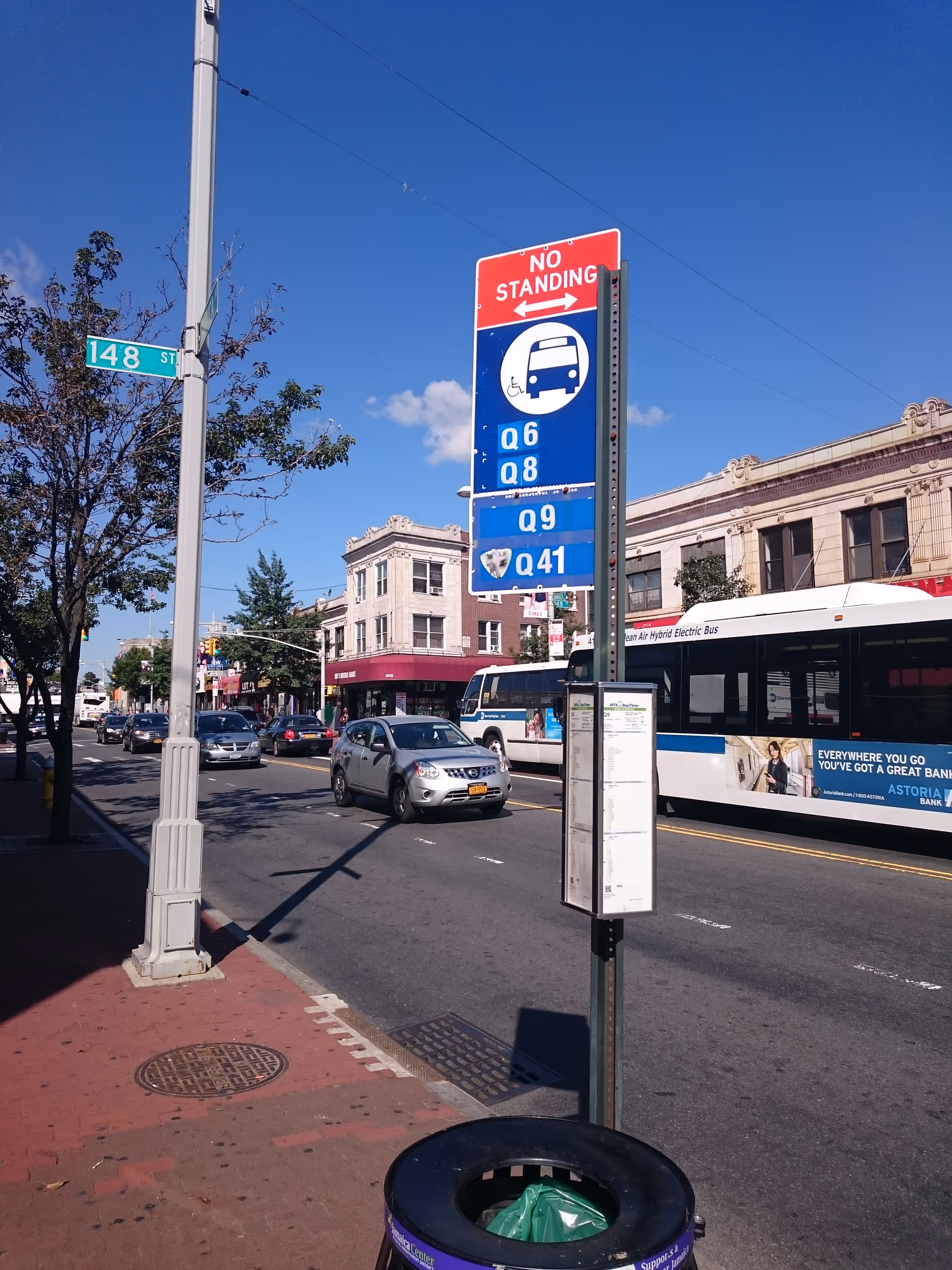 Busy city street with vehicles, a bus, storefronts, and a tall blue bus stop sign.